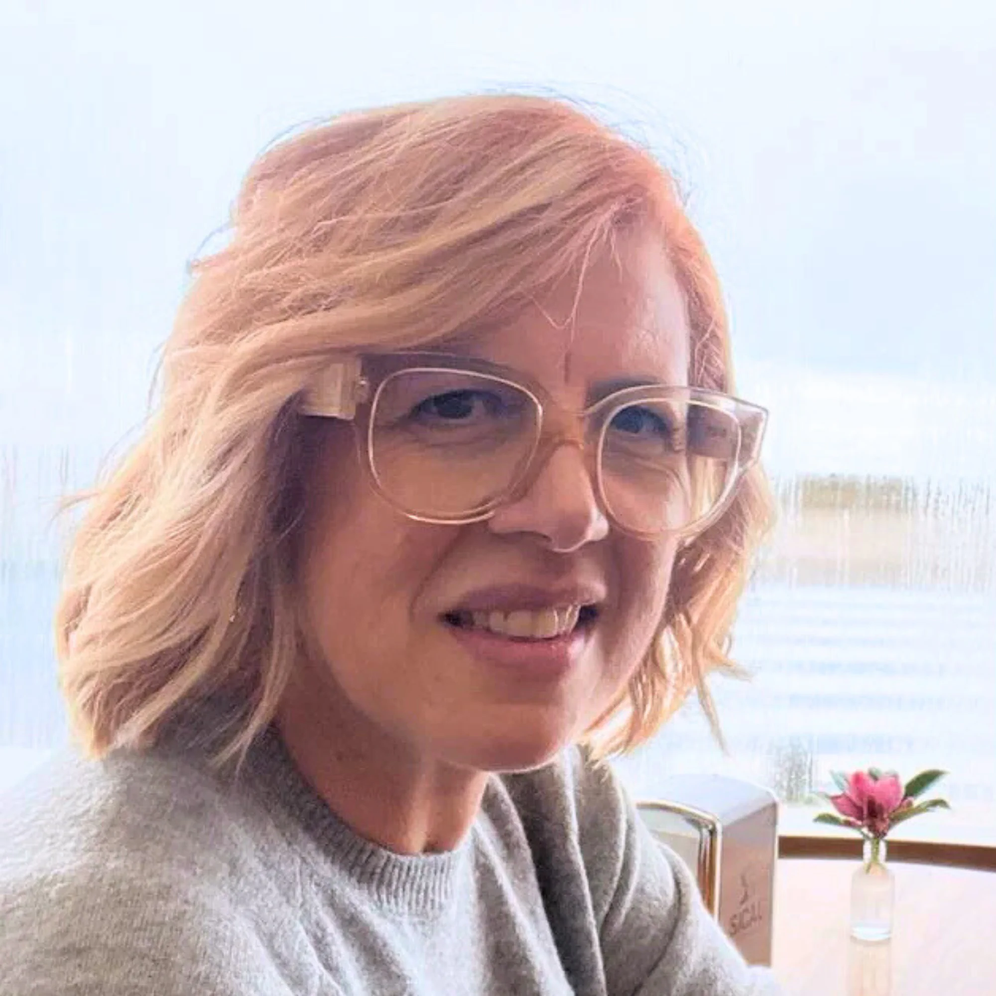Portrait d'une praticienne massage bien-etre Maria Nikita, cheveux blonds et lunettes, sourire doux pres d'une fenetre lumineuse face a l'eau